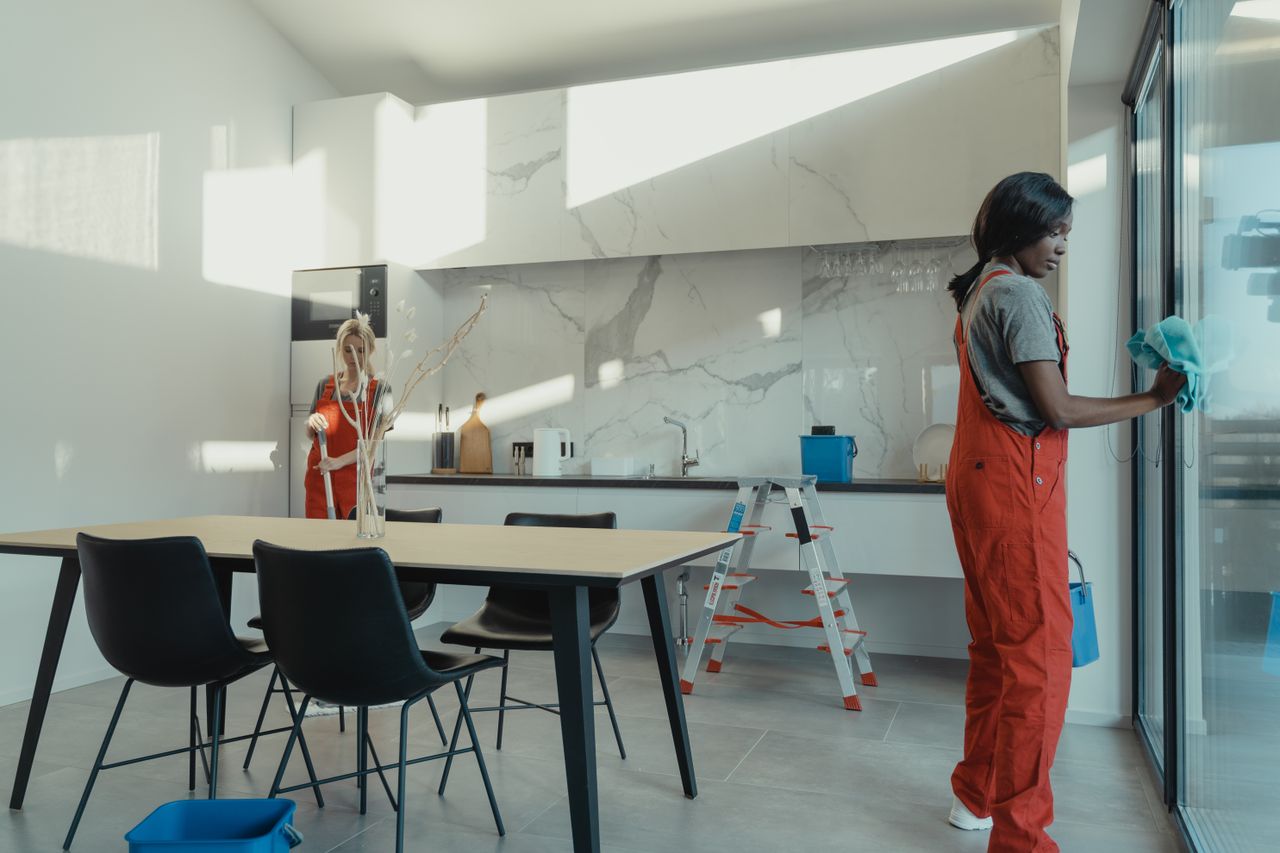 services-02 Two women in work clothes cleaning a kitchen with glass doors.