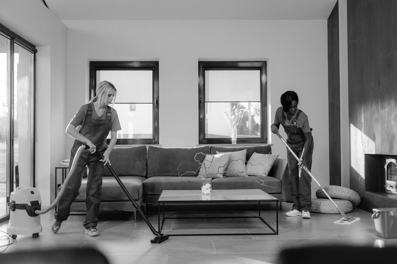 gallery-1 Two women in work clothes cleaning a modern living room using a vacuum and mop.